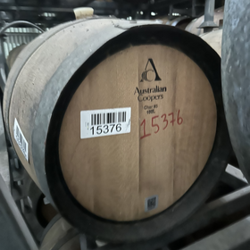 A wooden wine barrel with black metal hoops, showing the branded Australian Coopers end with a barcode sticker and red handwritten numbering, stored on metal racks.