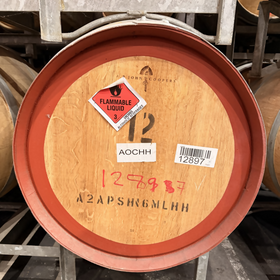 A large wooden whiskey barrel with a reddish-brown stave rim and natural wood interior, photographed on a metal rack in a warehouse. Stickers on the barrel include a red flammable liquid hazard label, white inventory tags, and handwritten red numbers.