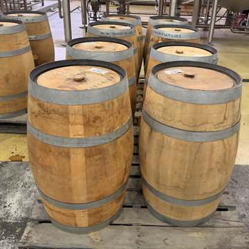 Several wooden whiskey barrels with metal bands sit on wooden pallets in a brewery or distillery setting, with stainless steel tanks visible in the background.