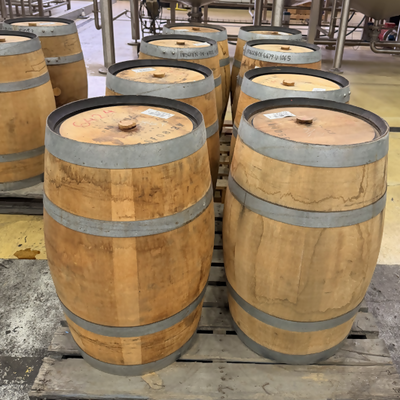 Several wooden whiskey barrels with metal bands sit on wooden pallets in a brewery or distillery setting, with stainless steel tanks visible in the background.
