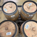 Four wooden wine barrels arranged in a 2x2 grid on a pallet in what appears to be a warehouse or storage facility. Each barrel has a metal band, a wooden top, handwritten markings, and a white barcode label.