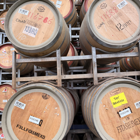 Multiple wooden wine barrels stacked on metal racks in a warehouse. The barrels have metal bands with handwritten numbers and labels, including one visible 'F3ASHMEX' stamp.